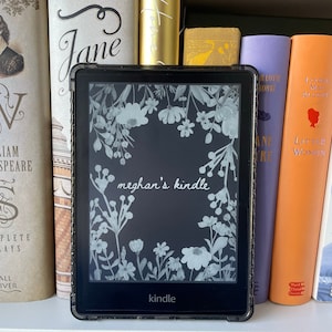 May include: A black Kindle e-reader with a floral design on the screen. The text "meghan's kindle" is displayed on the screen. The Kindle is surrounded by books on a shelf.