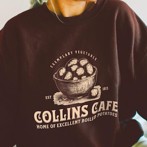 May include: Dark chocolate-colored sweatshirt with a vintage-style graphic. The graphic features a bowl of potatoes, the words "Exemplary Vegetable," and "Collins Cafe, Home of Excellent Boiled Potatoes." The sweatshirt has a crew neck.