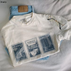 May include: An ivory-colored t-shirt with three blue postage stamp designs. The stamps feature architectural and landscape illustrations with text including "Netherfield Park", "Pemberley House & Woods", and "Longbourn House".