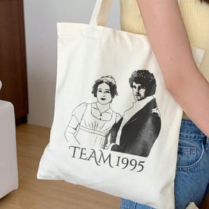 May include: White canvas tote bag with a black and white illustration of two people and the text "TEAM 1995".