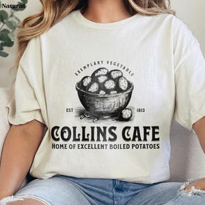 May include: A white t-shirt with a black graphic design of a bowl of potatoes. The text reads "Exemplary Vegetable Est 1813 Collins Cafe Home of Excellent Boiled Potatoes".