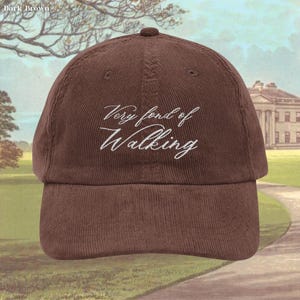 May include: Dark brown corduroy baseball cap with the embroidered text "Very fond of Walking" in white script. The cap has a curved brim and a rounded crown. The background shows a landscape painting.