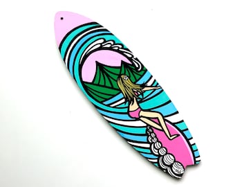 Puede incluir: Una tabla de surf rosa y azul con una escena de montaña verde y un surfista montando una ola. La tabla de surf tiene un contorno blanco y el texto "Mel" está escrito en la parte inferior.