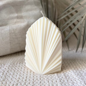 May include: Ivory-coloured, geometric candle with a fan-like design. The candle has a textured surface with vertical lines radiating from the base. The candle is unscented and stands on a textured, off-white surface.