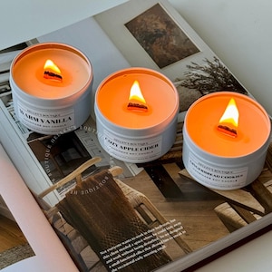May include: Three lit candles in light blue tins with wooden wicks. The candles have orange wax and are labelled "Warm Vanilla", "Cozy Apple Cider", and "Gingerbread Cookies". The candles are on a magazine with text and a wooden hanger.