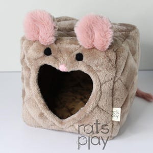 May include: A soft, brown pet house in the shape of a cube, designed to look like a mouse. It has a heart-shaped entrance, pink ears, black eyes, and a pink nose. A small, pink tail extends from the back. The text "rats play" is visible.
