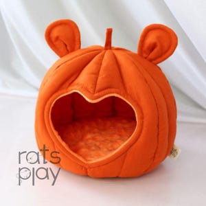 Puede incluir: Cama para mascotas con forma de calabaza naranja con una entrada en forma de corazón. La cama tiene dos protuberancias en forma de oreja y un tallo en la parte superior. El interior es de color naranja claro. El texto "rats play" es visible en la parte inferior izquierda.