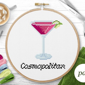 May include: Cross stitch pattern for a Cosmopolitan cocktail in a martini glass with a lime garnish. The pattern is in a wooden embroidery hoop and the text "Cosmopolitan" is below the image.