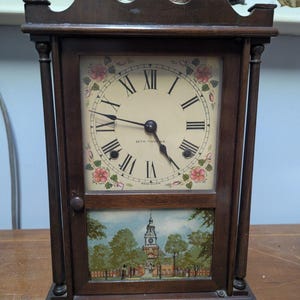 May include: A dark wood Seth Thomas clock with a cream-colored face and black Roman numerals. The clock face has floral accents and the words "Seth Thomas" are visible. The bottom panel features a landscape painting. The clock is Made in USA.
