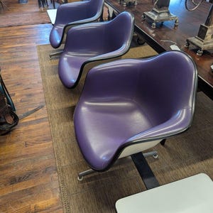 May include: Three purple armchairs with black trim and chrome legs are arranged on a brown rug. The chairs have a modern design with a curved seat and back. The background includes a wooden floor and antique furniture.