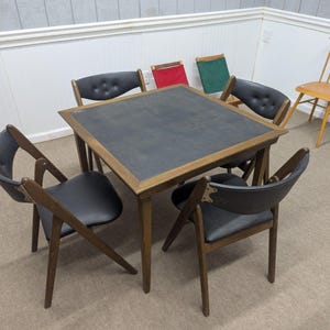 May include: A vintage square card table with a dark surface and wooden frame, surrounded by four black vinyl chairs. Additional chairs with red and green fabric backs are visible in the background. The set is on a neutral carpet.