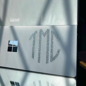 Può includere: Un coperchio per computer portatile color argento con una decalcomania monogramma argento glitterato che mostra le lettere 1MC in stile circolare, posizionato accanto a un piccolo emblema quadrato a quattro pannelli nero e argento.