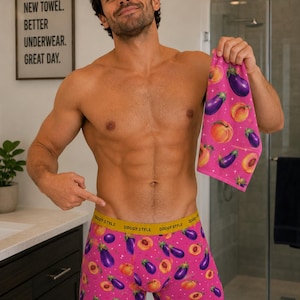 May include: Pink boxer shorts and a matching towel, both patterned with eggplants and peaches. The waistband of the shorts reads "DOGGY STYLE" in yellow. The towel is held up, and the shorts are being pointed at. The text on the wall says "NEW TOWEL. BETTER UNDERWEAR. GREAT DAY."