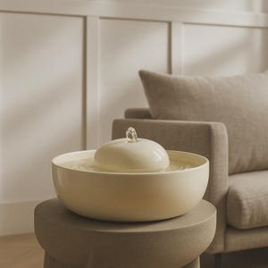 Könnte beinhalten: Ein cremefarbener Indoor-Wasserbrunnen mit einer abgerundeten Schale und einem zentralen, kuppelförmigen Element. Ein kleiner Ausguss an der Spitze der Kuppel gibt einen Wasserstrahl ab. Der Brunnen steht auf einem neutralfarbenen, runden Hocker, im Hintergrund ein Sofa.