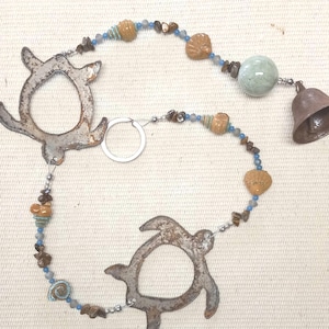 May include: A rustic wind chime featuring two metal turtle silhouettes, strung with beads in shades of blue, brown, and tan. A small bell hangs at the end. The chime is designed for outdoor decor.