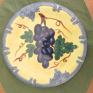 May include: A round, hand-painted ceramic plate with a yellow center and a blue and white patterned border. The plate features a cluster of dark purple grapes, green leaves, and brown vines.