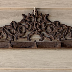 May include: A dark brown cast iron wall-mounted hook rack featuring an ornate, symmetrical scrollwork design along the top edge. The horizontal bar includes five evenly spaced hooks for hanging items, with visible screw holes for mounting.