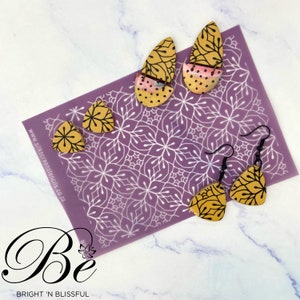 May include: A selection of handcrafted earrings in different forms, showcasing a mustard yellow hue with black floral patterns. The earrings are presented on a purple stencil with a floral design. The image also features the text "Be" and "BRIGHT 'N BLISSFUL".