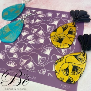 Könnte beinhalten: Ein Paar goldene und schwarze tropfenförmige Ohrringe mit einem Ginkgoblatt-Muster. Die Ohrringe befinden sich auf einem violetten Hintergrund mit einem weißen Ginkgoblatt-Muster. Der Text "BB55F" ist in der unteren rechten Ecke des Bildes sichtbar.