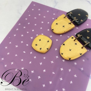 Peut inclure: Une paire de boucles d'oreilles dorées et noires avec un motif géométrique. Les boucles d'oreilles sont sur un fond violet avec un motif de croix blanches.