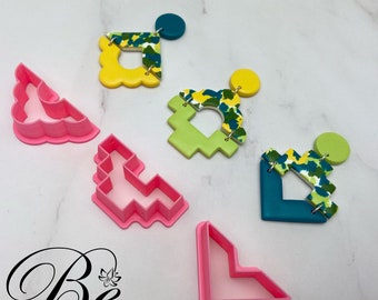 Puede incluir: Cinco cortadores de galletas de plástico rosa en diferentes formas: una forma curva, un ángulo recto, un ángulo izquierdo, una forma pixelada y una forma cuadrada con esquinas redondeadas. Los cortadores de galletas están sobre una superficie blanca.