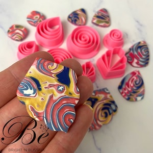 May include: A set of pink polymer clay cutters in various shapes, including a leaf, a flower, a swirl, and a fan. The cutters are arranged on a white surface with several pieces of colourful marbled polymer clay.