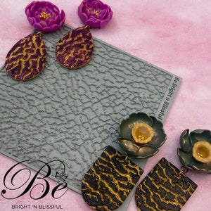 May include: Two pairs of earrings featuring a flower charm and a textured teardrop or rectangular shape. The flowers are purple and green with gold accents. The teardrop and rectangular shapes are purple and black with gold accents. The earrings are displayed on a gray textured surface with the text "Be Bright 'n Blissful".