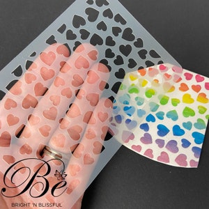 May include: A clear plastic stencil with a repeating pattern of small hearts. The stencil is being held over a rainbow-colored card with a similar heart pattern. The card has the text "Be Bright 'n Blissful" printed on the bottom.