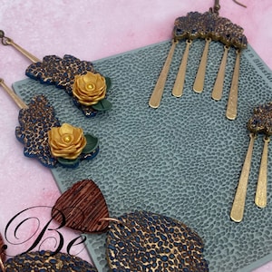 May include: Artisan-crafted earrings in various designs. One pair features a blue and gold leopard print with a gold flower and green leaves. Another pair has a blue and gold cloud-like top with gold dangling bars. The earrings are on a textured gray surface.