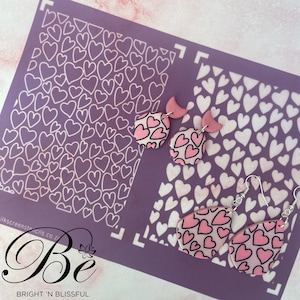 May include: Two pairs of pink heart-shaped earrings with a white heart pattern. The earrings are on a purple background with a stencil of white hearts.