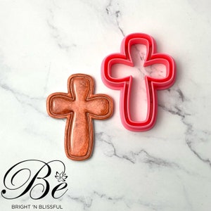 Könnte beinhalten: Ein rosa Plastik-Ausstecher in Form eines Kreuzes und ein braunes Kreuz-Kekse. Der Ausstecher ist etwas größer als der Keks.