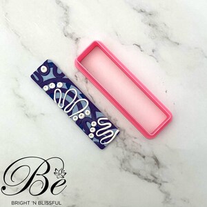 Op de afbeelding: Een roze plastic rechthoekige koekjesvormer met een blauwe en witte gepatroneerde polymer clay-staaf erin. De staaf heeft een wit swirl-ontwerp met witte stippen.