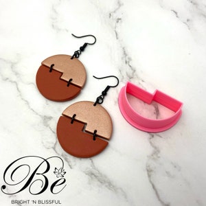 May include: A pair of earrings with a copper and gold color scheme. The earrings are made of two half circles that are joined together with a black metal connector. The earrings are hanging from black metal hooks. A pink plastic mold is also shown in the image.