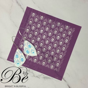 May include: A pair of white teardrop-shaped earrings with a blue and white peacock feather design. The earrings are next to a purple stencil with a white peacock feather pattern.
