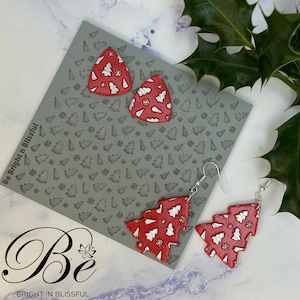 May include: A pair of red and silver Christmas tree earrings with a white snowflake pattern. The earrings are on a gray background with a pattern of small Christmas trees.