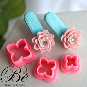 Puede incluir: Dos pendientes de suculentas de arcilla polimérica de color rosa con un fondo azul turquesa. Los pendientes están hechos con un pequeño molde en forma de flor y una pedrería en el centro. La imagen también muestra dos moldes de plástico rosa para hacer los pendientes de suculentas.
