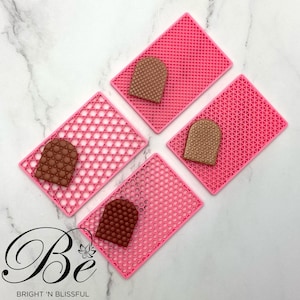 May include: Four pink silicone baking mats with a grid pattern. Each mat has a brown chocolate-like object in the shape of a heart on top.