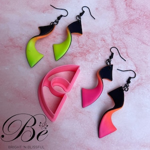 May include: Three pairs of colorful polymer clay earrings with black metal hooks. The earrings are shaped like a curved line with a gradient of color. The earrings are displayed on a pink surface with a pink plastic mold.