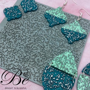 May include: A close-up of a silicone mold with a textured surface. The mold is used to create earrings with a teal and mint green design. The earrings are made of polymer clay and have a silver metallic finish. The text "Be Bright 'N Blissful" is visible in the bottom left corner of the image.