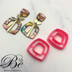 May include: Two pairs of colorful square earrings with gold accents and two pink plastic square cutters. The earrings have a marbled design with gold leaf accents. The cutters are designed to cut out square shapes from clay.