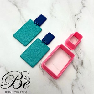 May include: Two pairs of rectangular polymer clay earrings with a blue and teal geometric pattern. The earrings are shown next to two pink plastic rectangle-shaped cookie cutters.