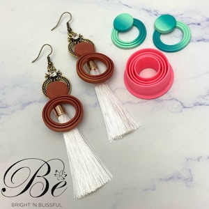 May include: A pair of earrings with brown and white tassels. The earrings feature a gold-tone metal setting with a clear rhinestone and a layered circle design. The circles are brown with a gradient effect. The earrings are displayed on a white marble surface with pink and green circle-shaped moulds.