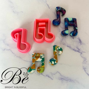 May include: Five music note shaped cookie cutters, two are pink, two are blue and purple with glitter, and one is green and gold with glitter. The cookie cutters are on a white marble surface. The text "Be" is written in black with a pink lotus flower below it. The text "BRIGHT 'N BLISSFUL" is written in black below the lotus flower.