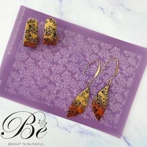 May include: A pair of gold-toned earrings with a teardrop shape and a black and orange leaf pattern. The earrings are displayed on a purple background with a white leaf pattern.