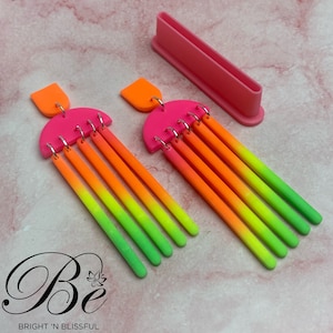 May include: A pair of neon-coloured earrings with a distinctive design. The earrings have a bright pink half-circle top with an orange rectangular accent. Below, multiple neon-coloured rods transition from orange to green. A pink rectangular tool is also present.