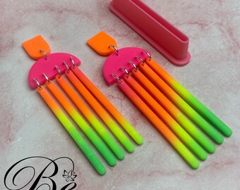May include: A pair of vibrant, neon-colored earrings with a unique design. The earrings feature a bright pink half-circle top with an orange rectangular accent. Below, multiple neon-colored rods transition from orange to green. A pink rectangular tool is also present.