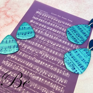 May include: A pair of teal blue teardrop-shaped earrings with a musical note pattern. The earrings are on a purple background with sheet music printed on it. The text on the sheet music reads "Love's Old Sweet Song".