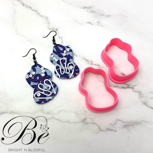 Può includere: Due paia di orecchini con motivo viola e bianco con ganci neri e due tagliapasta in plastica rosa a forma di orecchini.