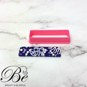Op de afbeelding: Een roze plastic rechthoekige koekjesuitsteker met een blauwe en witte gepatroneerde polymer clay-staaf erin. De staaf heeft witte swirls en stippen op een blauwe achtergrond. De tekst "Be" is zichtbaar in de linkerbenedenhoek van de afbeelding.