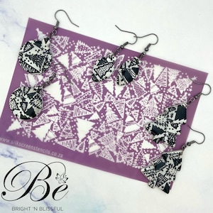 May include: A set of three pairs of black and white patterned earrings in the shape of a circle, a teardrop, and a Christmas tree. The earrings are on a purple background with a white pattern of Christmas trees.